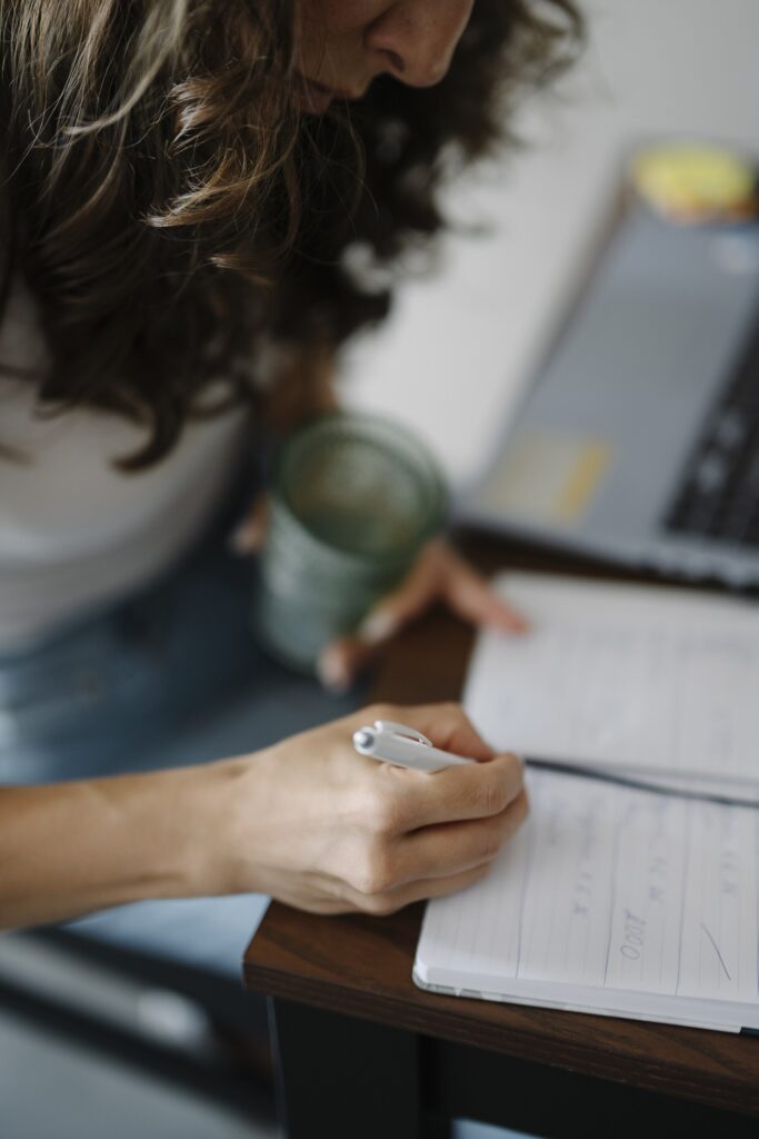 Person writes in notebook while holding drink near laptop